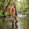 PenBag PreparedDad wading a forest stream with the PBS 125-in-1 survival kit — orange tactical pack and first-aid pouch in hand